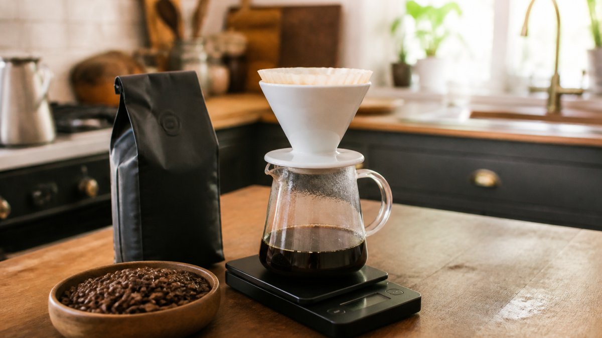 A home coffee brewing setup on a kitchen counter with a pour-over dripper, a bag of whole bean coffee, and a digital scale, shot in warm natural light
