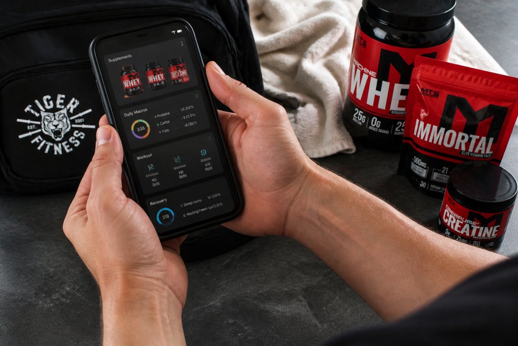 Close-up of hands holding smartphone with fitness app dashboard showing supplement stack, daily macros, workout metrics, and recovery data on bright screen
