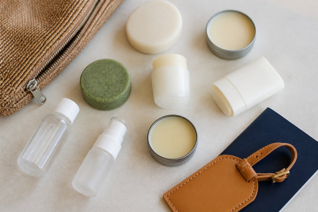 Flat lay of compact travel beauty products including solid shampoo bar, solid deodorant, mineral sunscreen, solid perfume, and small refillable containers arranged on a light surface with a travel passport and luggage tag