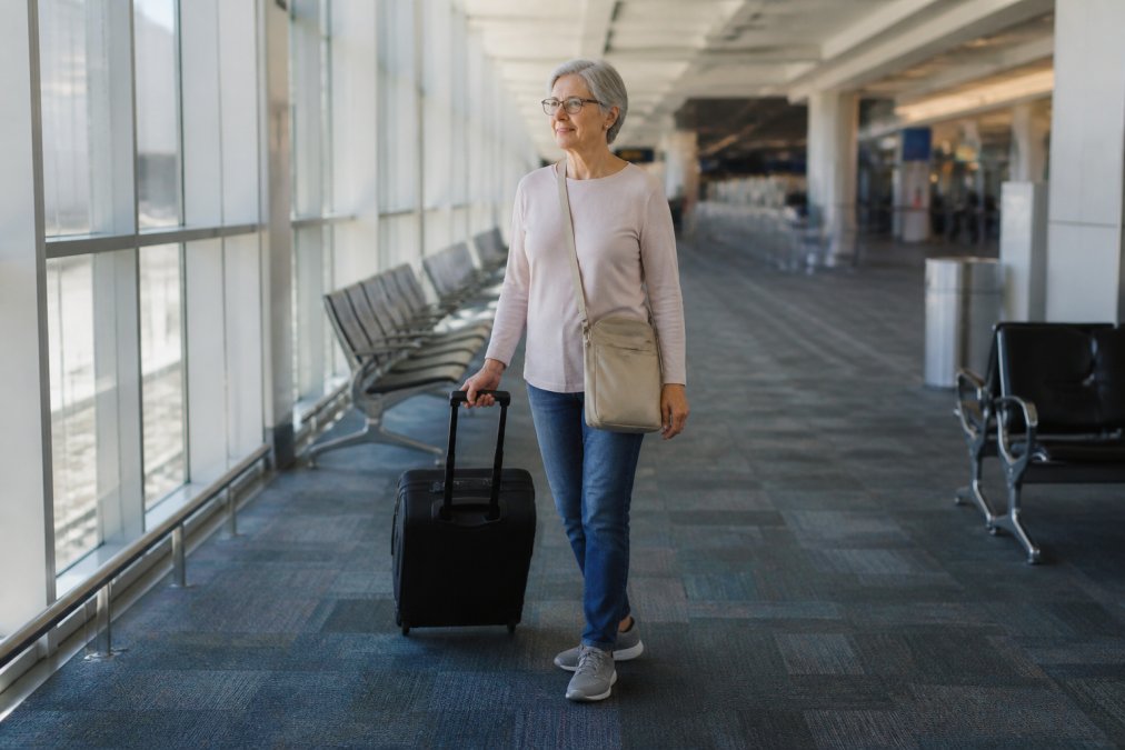 A traveler with a rolling suitcase wearing comfortable wide-width walking sneakers in a bright airport terminal, looking relaxed and ready to board