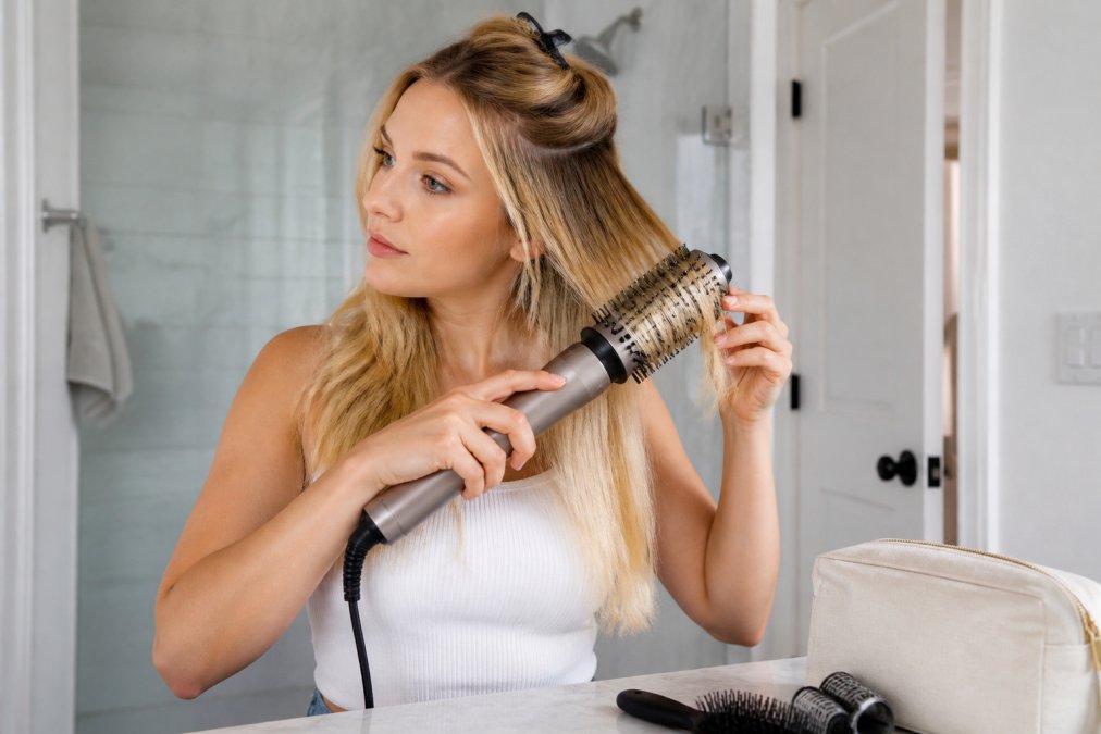 Woman with fine blonde hair using the best travel hair dryer brush for fine hair to curl a section at the roots.