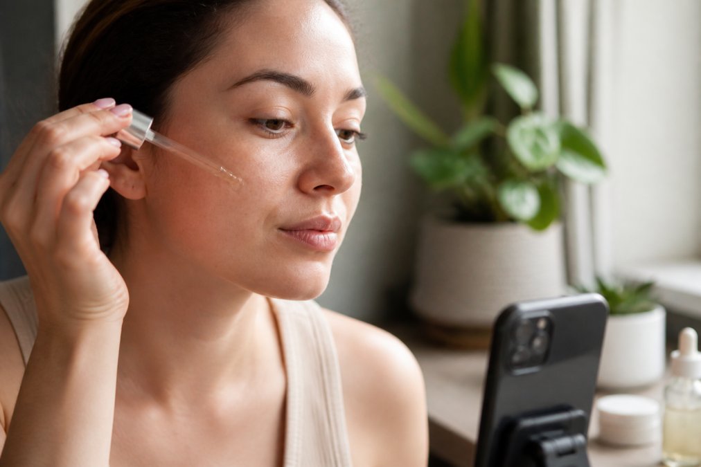 A close-up of a woman applying a hydrating serum to her face, with a smartphone showing a TikTok beauty video in the background, natural light