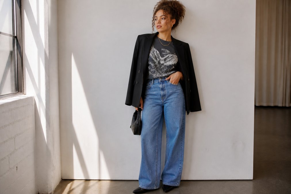 A young adult with a bold graphic tee half-tucked into high-waisted wide-leg jeans, a structured blazer draped over their shoulders, standing in front of a bright white wall with soft natural window light, relaxed confident posture