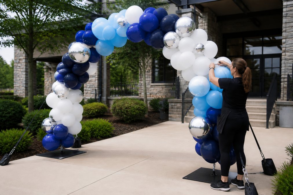Professional balloon artist installing custom foil and latex balloon arch at outdoor venue with proper anchoring systems visible, showing structural support and tension in bright daylight