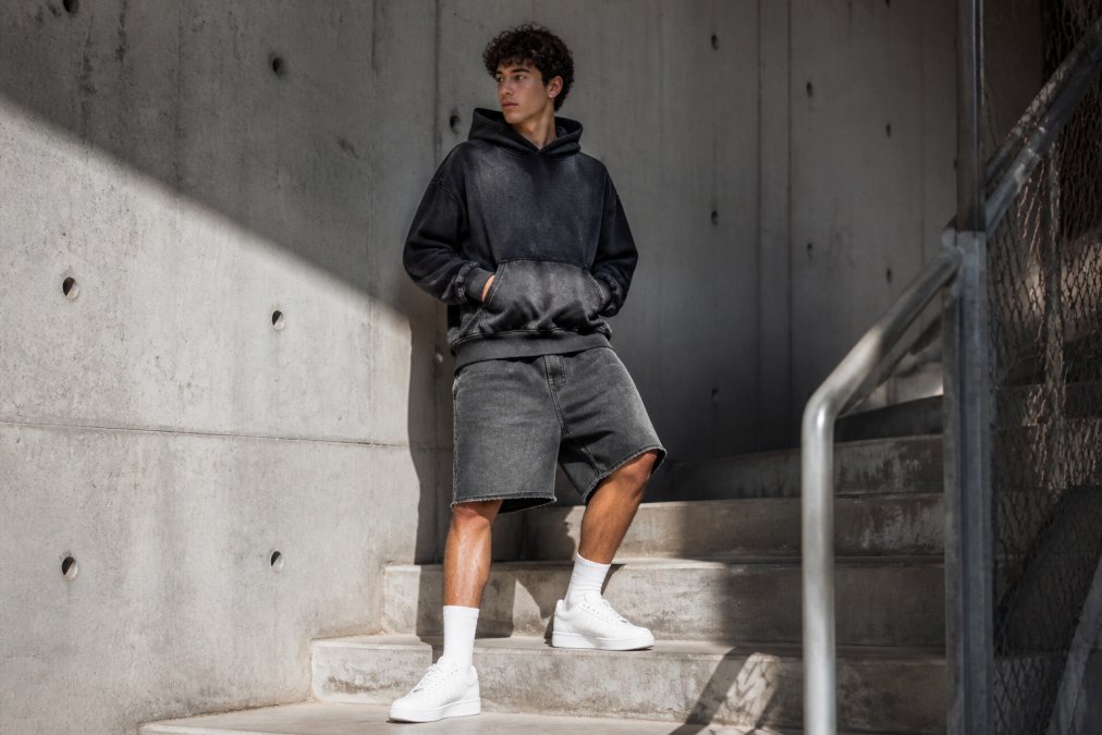 A person wearing an oversized, deeply washed cotton hoodie with a drop shoulder and relaxed-fit distressed shorts, standing on a concrete staircase in natural daylight with clean white sneakers