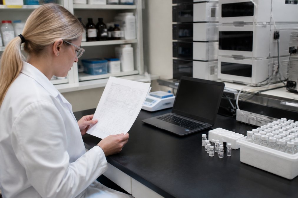 A researcher in a lab coat examining a printed certificate of analysis document at a laboratory bench, with a laptop and HPLC equipment visible in the background under fluorescent lighting