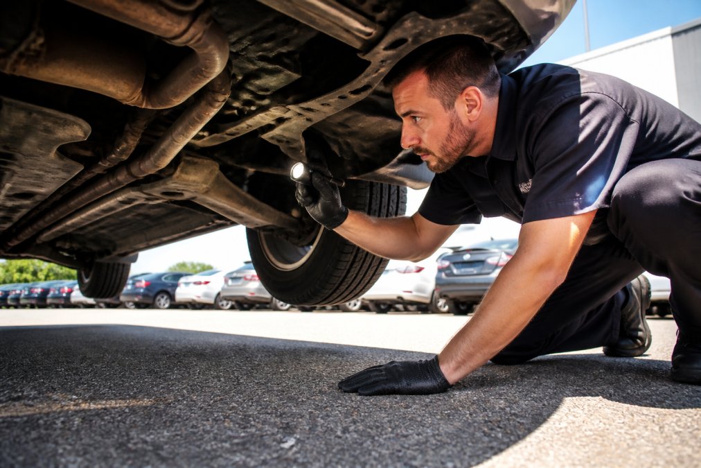 Professional mechanic crouching beside a used sedan, using a flashlight to inspect the undercarriage for rust and corrosion on a sunny dealership lot