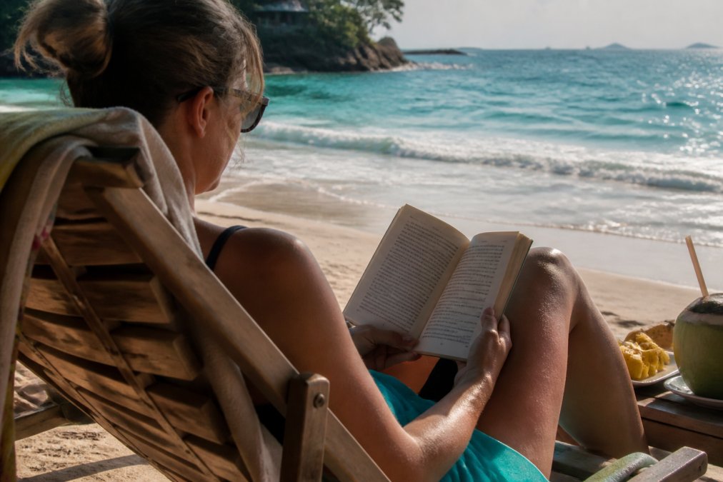 Reader relaxing on a beach chair with an open mystery novel, tropical waves visible in the background, golden afternoon sunlight casting shadows across the pages