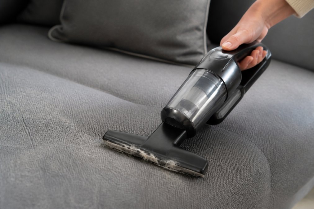 Handheld vacuum with a rubber attachment removing pet hair from a gray fabric sofa during upholstery cleaning.