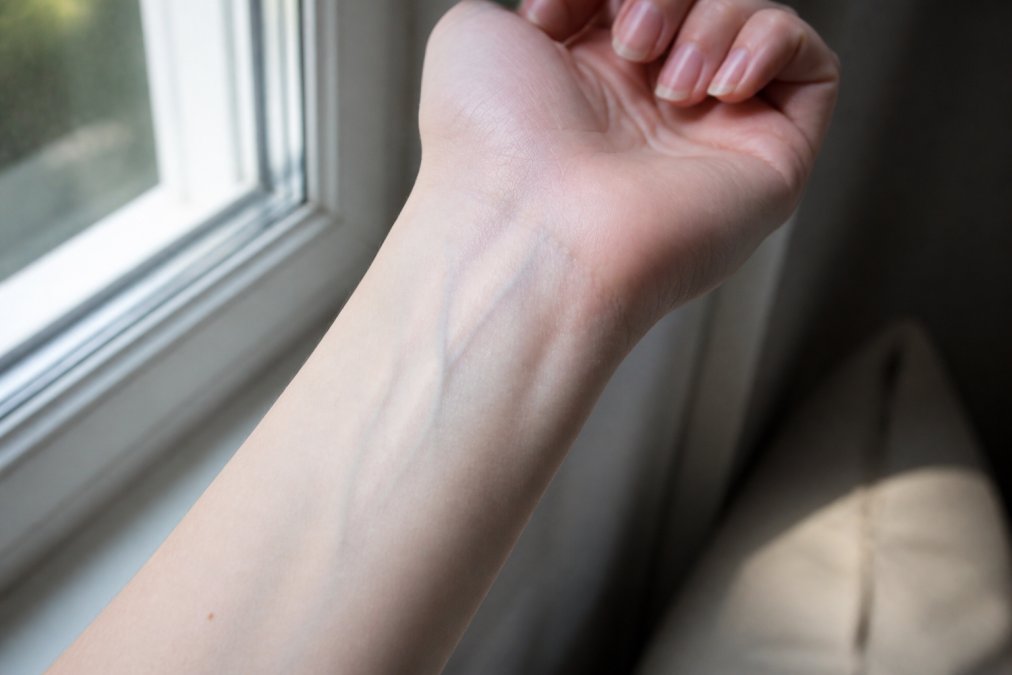 Close-up of a woman's inner wrist in natural daylight near a window, showing veins clearly visible through fair skin, with soft natural light illuminating the skin to demonstrate the vein color test for undertone identification