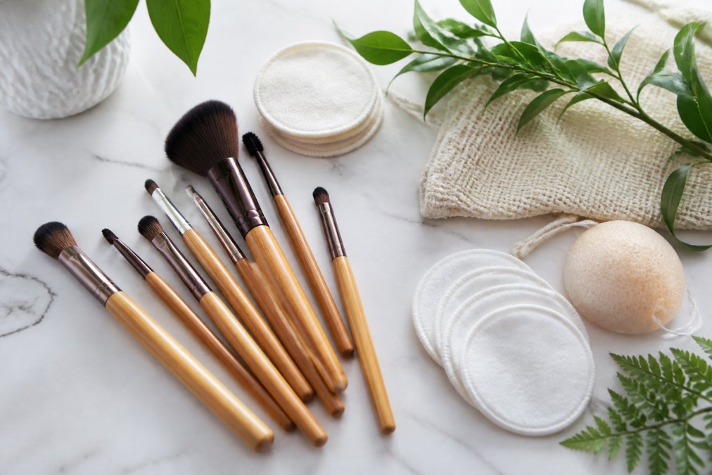Flat lay of soft makeup brushes with bamboo handles, reusable cotton pads, and gentle cleansing tools arranged neatly on a light marble surface with green botanical accents
