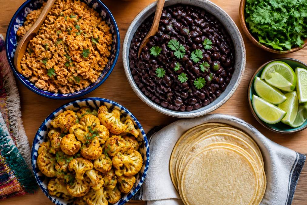 Overhead shot of a vibrant taco bar spread with colorful bowls of cooked black beans, seasoned crumbled tofu, sautéed cauliflower, fresh cilantro, lime wedges, and corn tortillas arranged on a wooden table