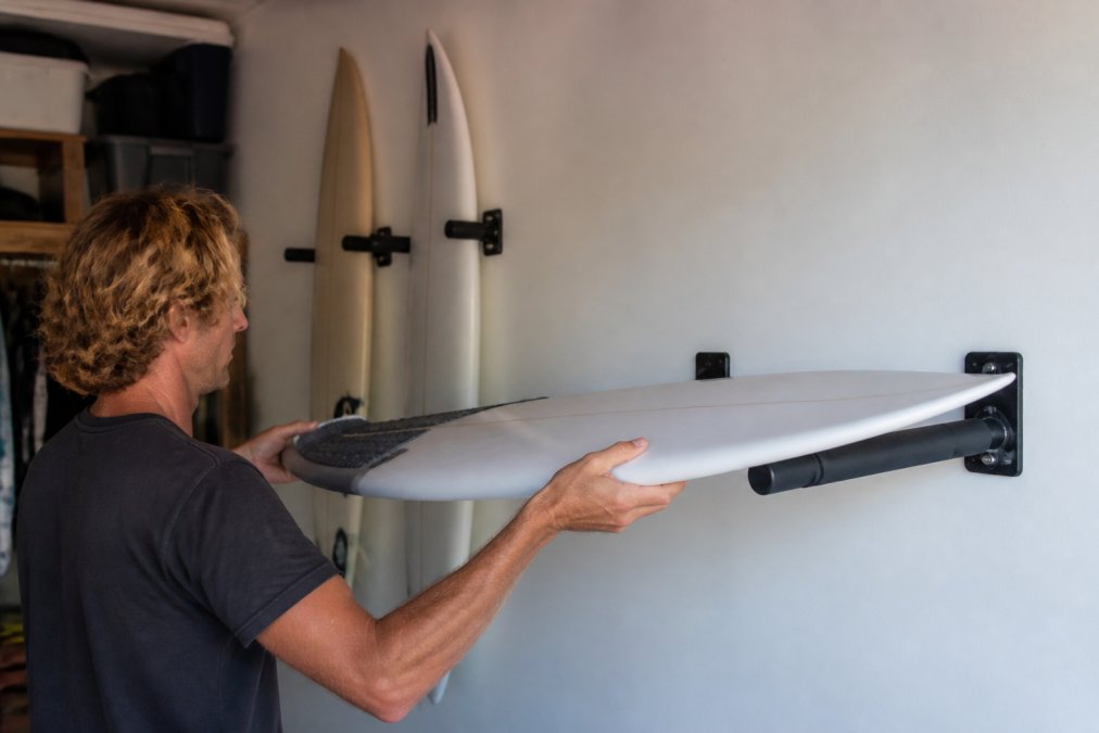 Surfer placing a shortboard into a foam-padded wall mount among other surfboard display racks in a home garage.