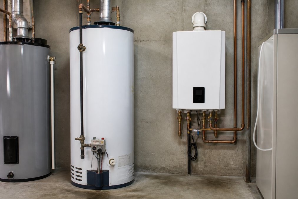 A row of modern water heaters in a bright basement utility room, a tall white gas tank unit beside a compact wall-mounted tankless model, showing the size difference between the two types