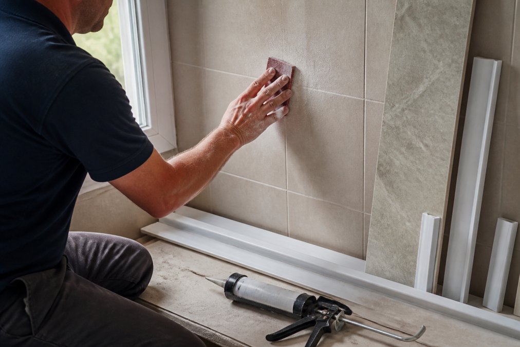 Professional tradesperson using coarse sandpaper on bathroom wall tiles, with dust particles visible in natural bathroom lighting, preparing surface for shower panel adhesive application