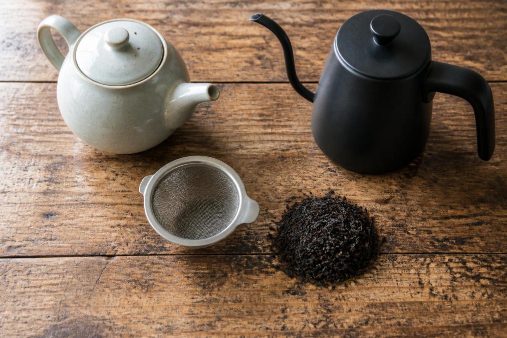 Flat lay of loose leaf tea brewing equipment on a worn wooden surface: a ceramic teapot, a round mesh infuser, a matte black tea kettle, and a small mound of dry dark loose leaf tea leaves, soft natural light from the left