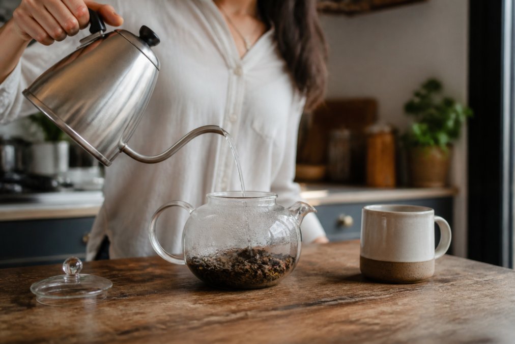 A person pouring hot water from a gooseneck kettle into a clear glass teapot filled with loose leaf tea, steam rising, on a wooden kitchen counter with a ceramic mug nearby