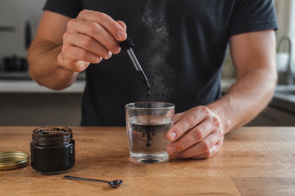 Close-up of a man's hands holding a small glass of warm water with a dropper of dark shilajit resin being squeezed into it, steam rising gently, on a wooden kitchen counter with a jar of resin nearby