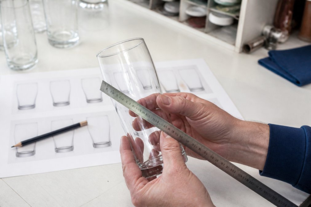 Close-up of hands holding a curved pint glass against a printed proof sheet on a bright workbench, checking logo position with a ruler