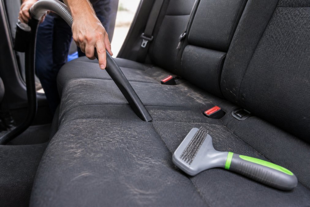 Person using a vacuum crevice tool to remove dog smell and hair from a car seat with a pet brush nearby