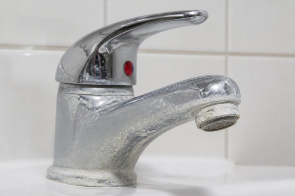 Close-up of a chrome bathroom faucet with thick white mineral deposits and limescale crust around the base and aerator, photographed under bright bathroom lighting against white tile