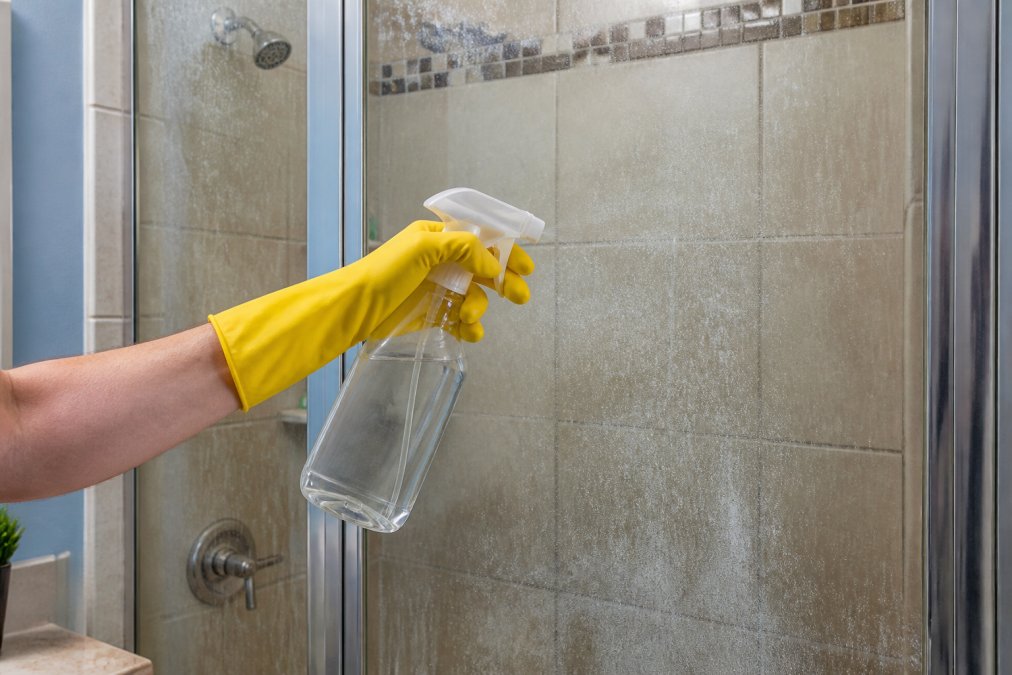 A person wearing yellow rubber gloves spraying white vinegar cleaning solution onto a glass shower door covered in cloudy white hard water mineral film, in a bright modern bathroom