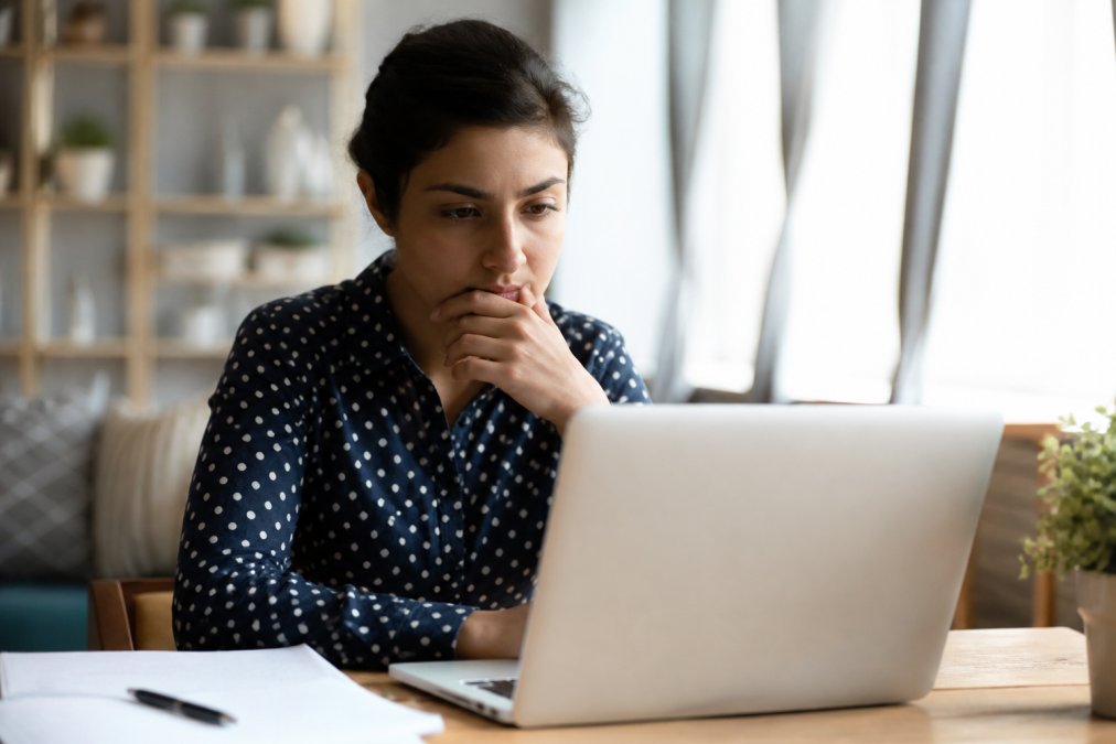 Person pausing before making an online purchase, looking thoughtful at a laptop with hand on chin, showing a moment of deliberation and hesitation