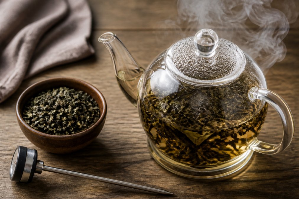 Loose tea leaves expand in a glass teapot as you brew loose leaf tea, with a digital thermometer on a wooden counter.