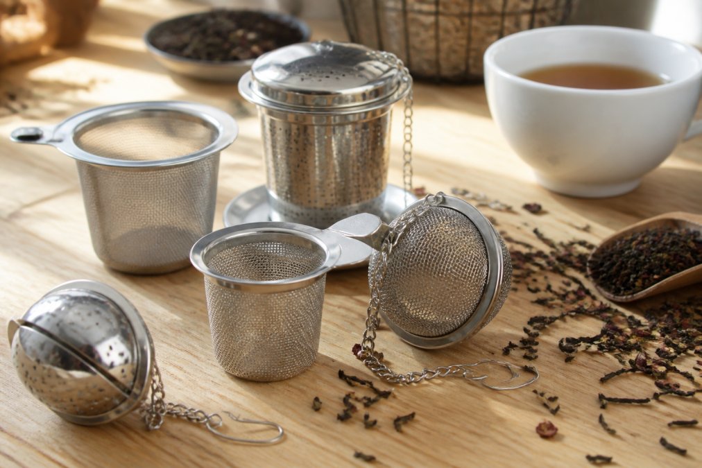 Collection of various stainless steel tea infusers, mesh strainers, tea balls, and infuser baskets arranged on a light wooden surface with loose tea leaves and a white ceramic cup nearby, natural afternoon lighting