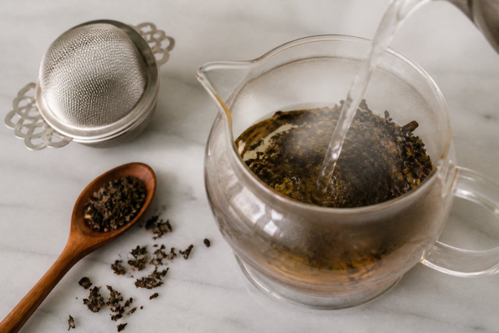Close-up overhead view of loose leaf tea steeping in a glass brewing vessel, hot water pouring over dark tea leaves, steam rising, with a stainless steel infuser and wooden spoon nearby on a light surface, warm natural lighting