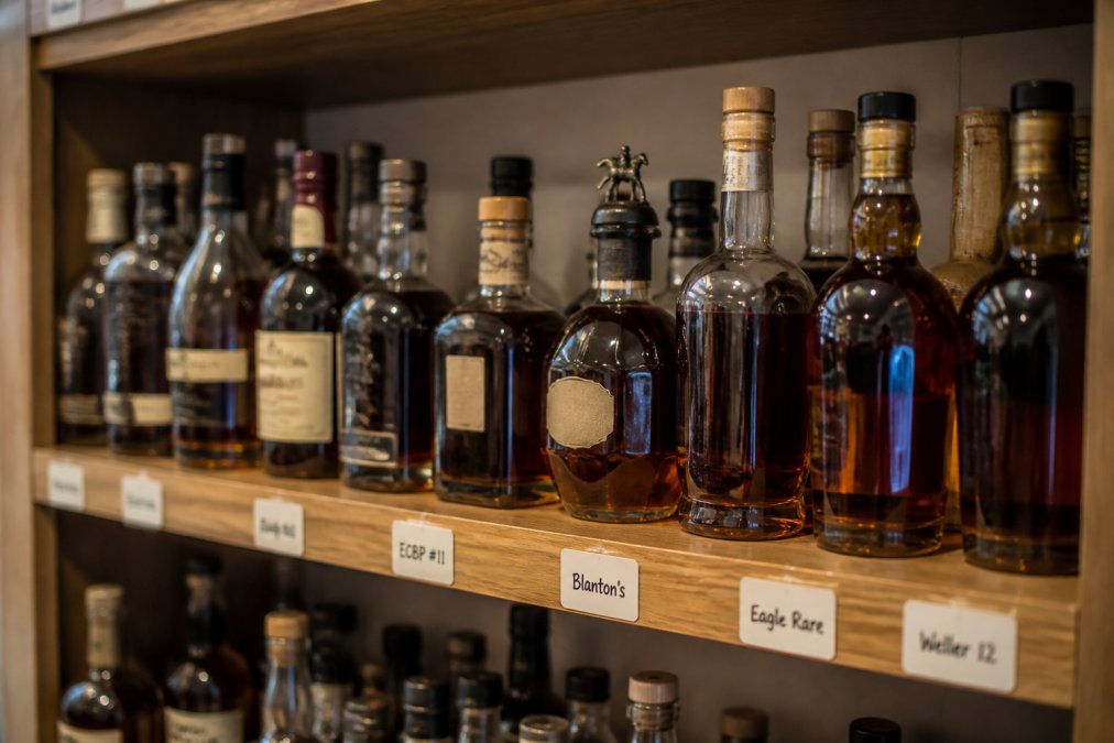 A well-organized home bar shelf with rows of bourbon bottles neatly arranged, some with handwritten tags on the shelf edge, warm ambient lighting