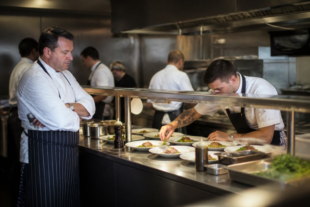 Head chef observing a candidate working at the pass during a busy kitchen service, with multiple dishes being plated and prepared in the background