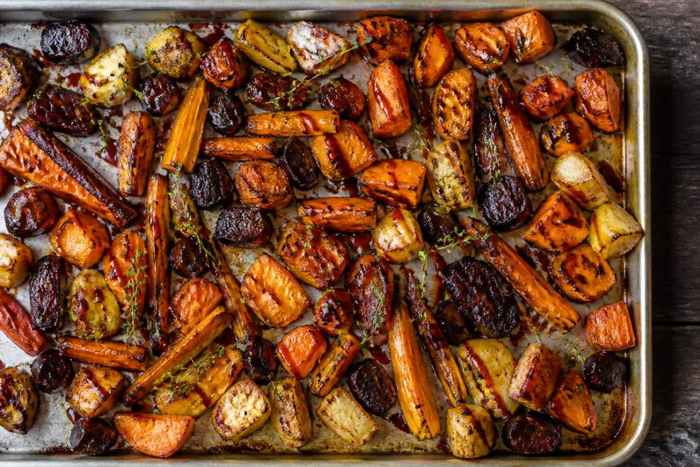 Overhead shot of a sheet pan with roasted root vegetables in rich caramel-brown tones, with a drizzle of spicy sauce visible and fresh thyme sprigs scattered on top