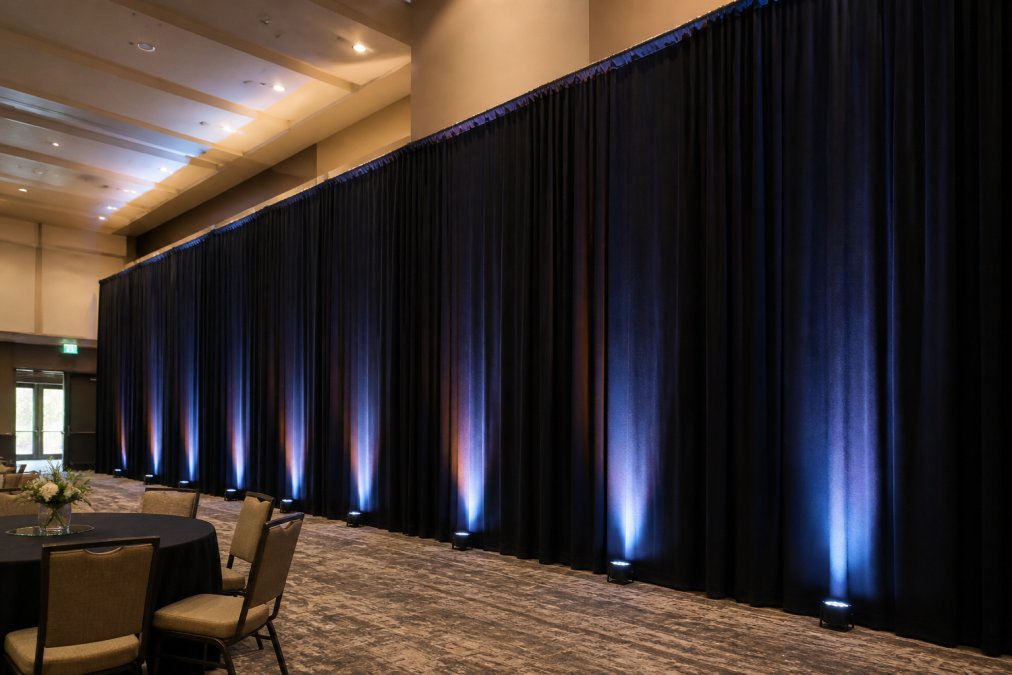 Professional event space with black pipe and drape system installed floor-to-ceiling showing proper tension and alignment, with uplighting creating ambient color wash against the draped fabric
