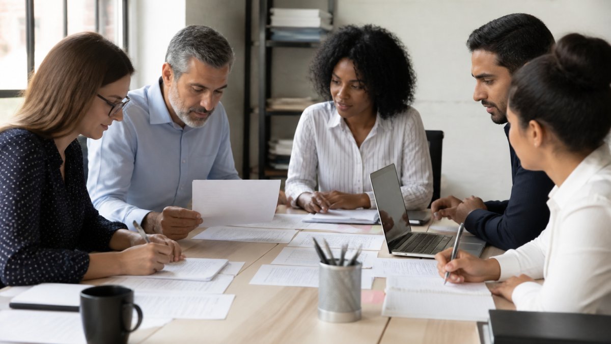 Professional team of diverse members gathered around a conference table reviewing printed project documents and taking notes, with a laptop open showing a shared workspace on screen in modern office with natural window lighting