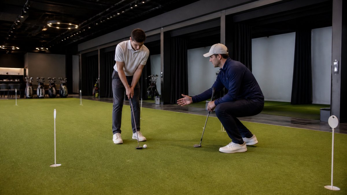 A golfer practicing a putting drill on a PuttView putting studio green, with a coach observing and providing feedback, simulator bays visible in the background of a premium indoor golf facility