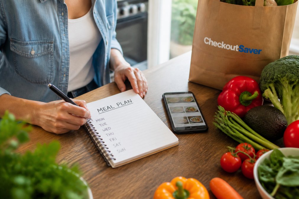 Person writing a weekly meal plan at a kitchen table with a notebook, fresh vegetables, and a smartphone showing a meal planning app nearby