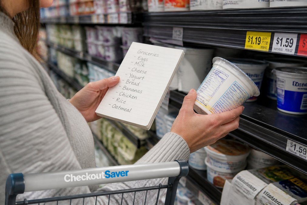 Shopper holding a handwritten grocery list while comparing product prices and checking unit prices on store shelves