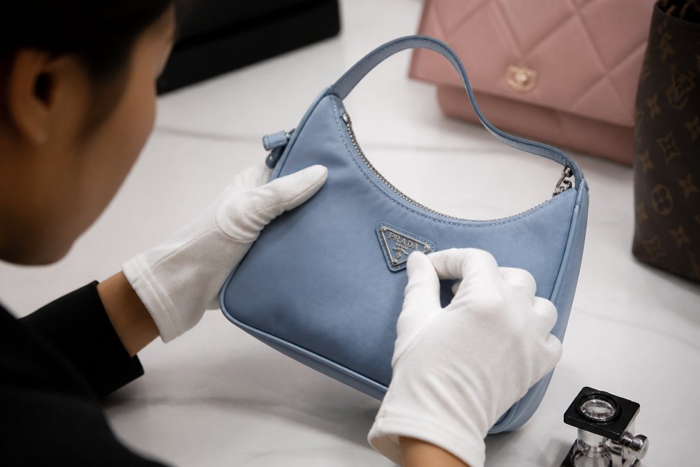 Close-up of gloved hands carefully examining the leather surface and logo hardware of a luxury designer handbag on a clean white surface in a well-lit authentication studio