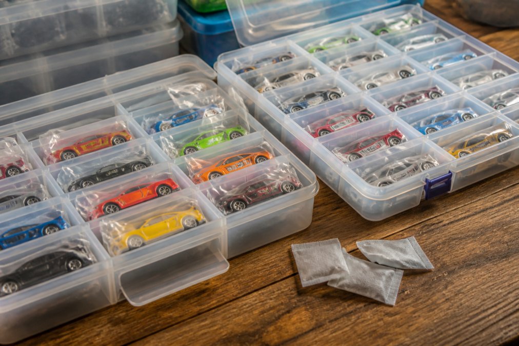 Close-up of organized compartmentalized storage bins containing Hot Wheels diecast cars in individual poly bags, arranged on a flat wooden surface under warm workshop lighting with small silica gel desiccant packets placed beside the open bins