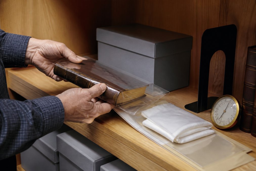 Close-up of hands carefully placing a leather-bound signed first edition into an acid-free archival Mylar sleeve, with a Solander box, bookends, and a hygrometer visible on a wooden shelf beside protective archival materials
