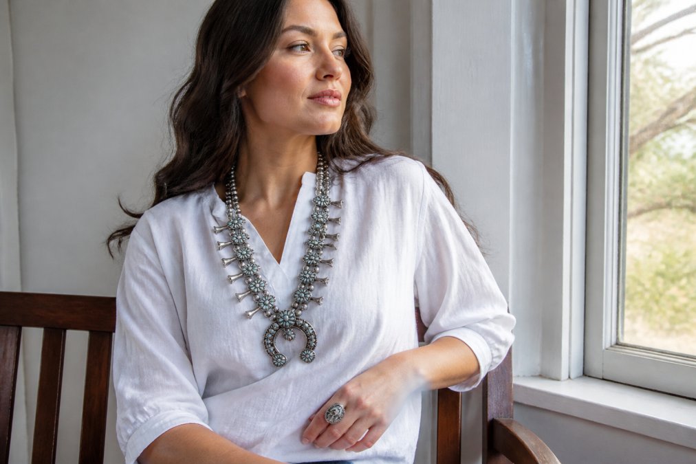 A woman wearing a large sterling silver squash blossom necklace over a simple white linen blouse, seated near a sunlit window with minimal other accessories visible