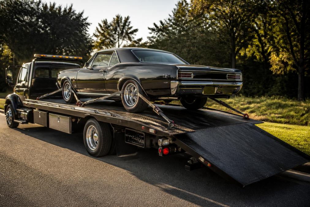 Professional flatbed tow truck with pristine 1960s classic car fully secured with soft-tie straps and hydraulic lift gate lowered, showing proper loading technique from the side in afternoon sunlight