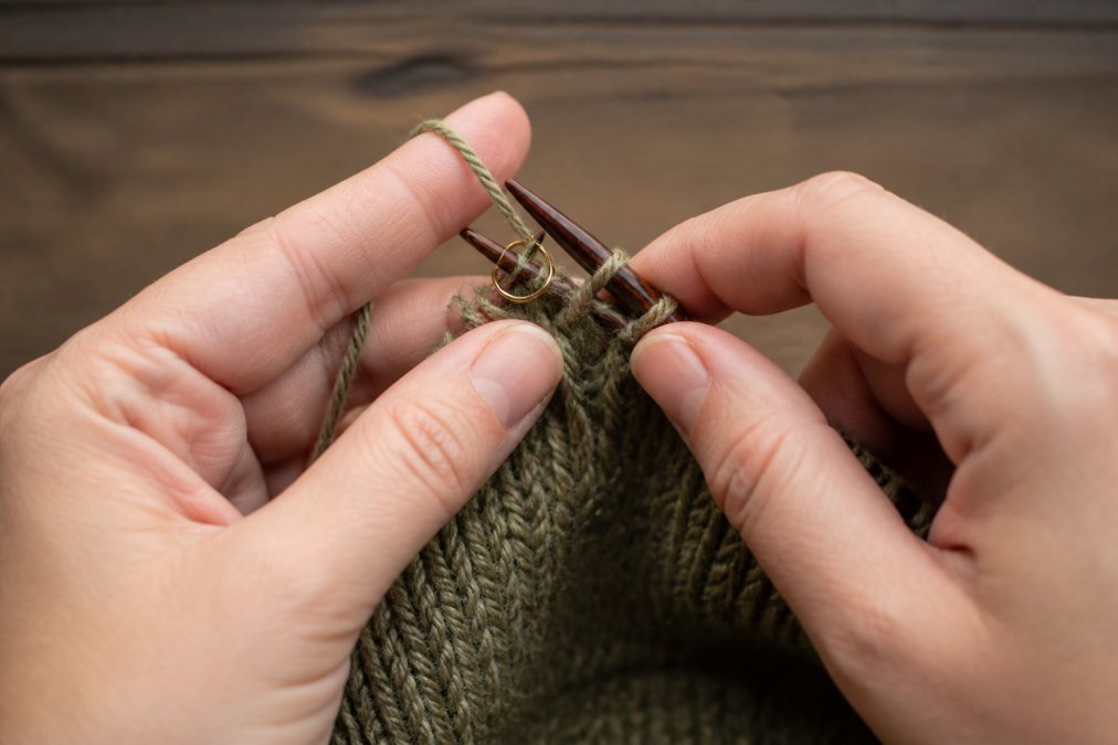 Close-up of hands placing a ring stitch marker onto a knitting needle mid-project, showing proper positioning between stitches with yarn in progress