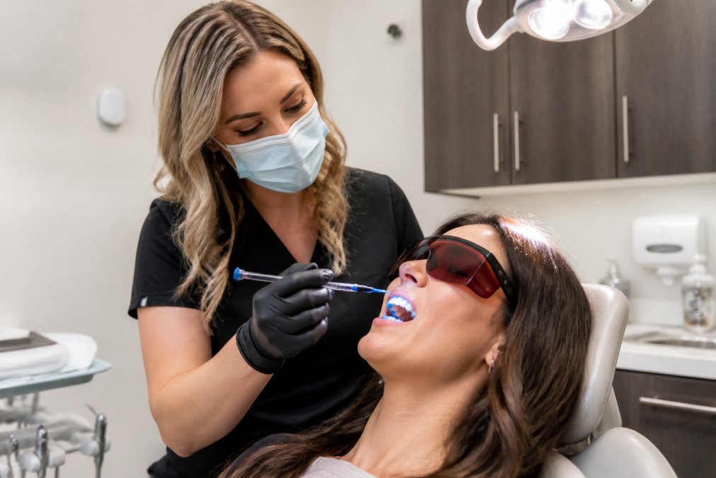 Professional dentist applying professional-grade whitening gel to patient's teeth in clinical chair under bright overhead dental light