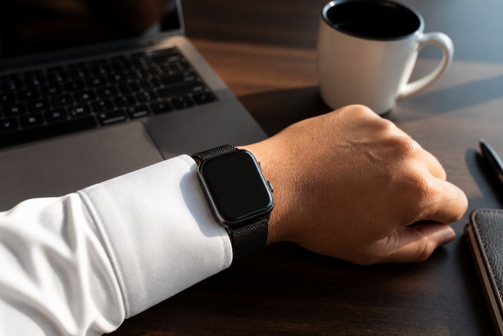 Close-up of a professional's wrist wearing a sleek graphite smartwatch over a white shirt cuff, with a laptop keyboard and coffee cup visible on a modern office desk in warm natural light