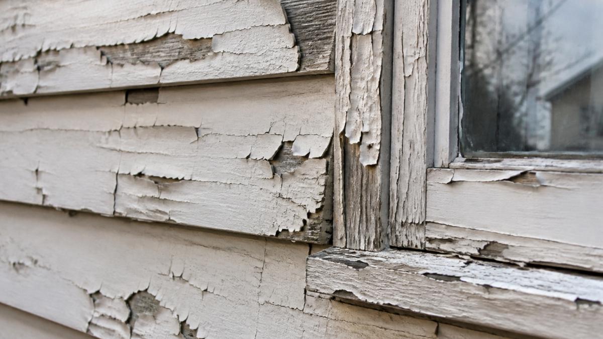 Close-up of peeling and cracked exterior paint on a wood-sided house, showing visible freeze-thaw damage and flaking layers near a window frame