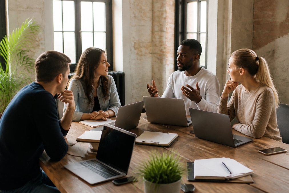 Team of developers collaborating around a table with laptops, discussing website infrastructure and scaling strategies in a modern office environment with natural lighting