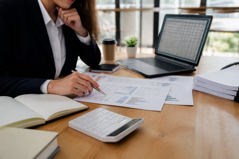 A business professional reviewing financial documents and a laptop with spreadsheets at a clean modern desk, calculator and notebook nearby