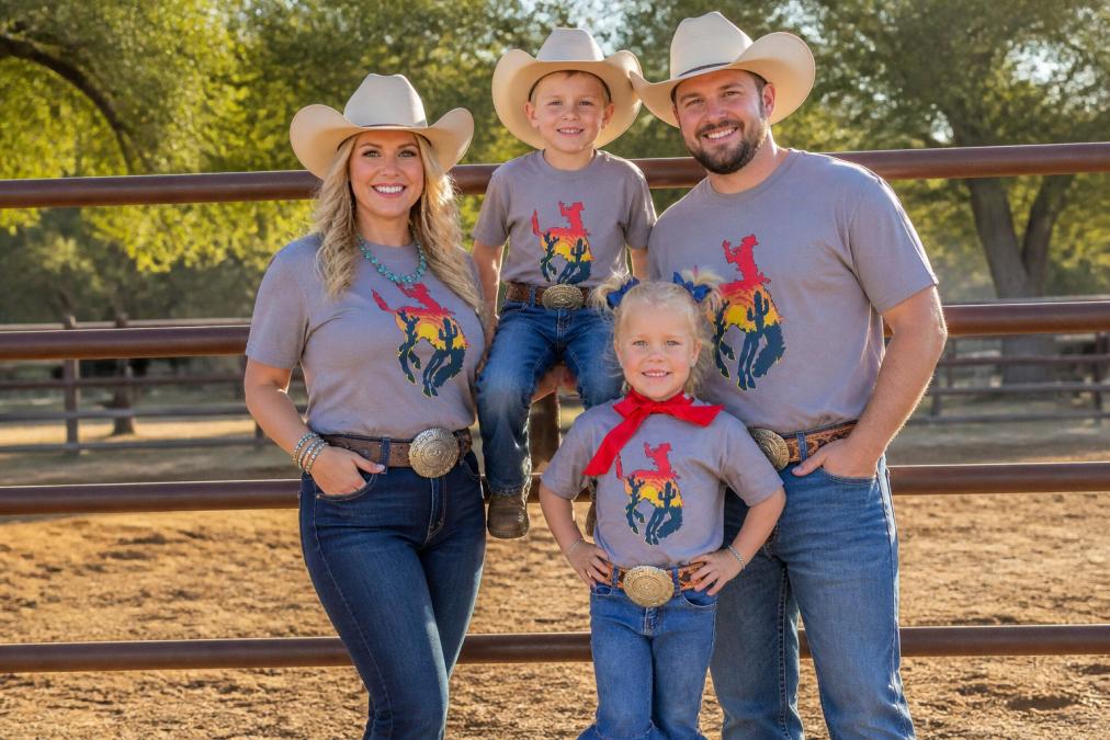 A family of four dressed in coordinated western wear, denim, graphic tees, and cowboy hats, standing together on a ranch fence in warm afternoon sunlight, smiling naturally
